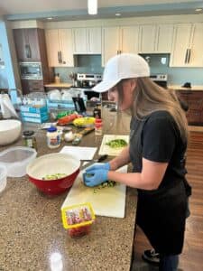 Person preparing food in a kitchen, chopping vegetables on a cutting board, with various ingredients on the counter.