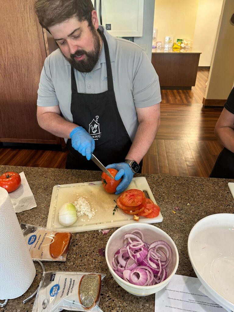 Man slicing tomatoes on a kitchen counter with chopped onions, spices, and red onion rings nearby.