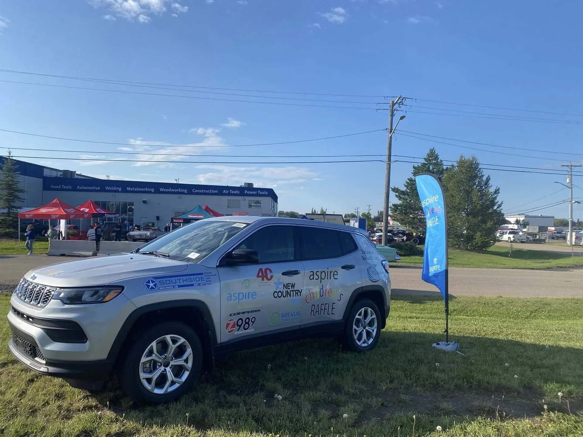 Jeep with promotional decals parked by a roadside event, with tents and a flag in the background.