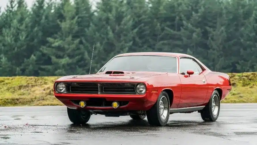 Classic red muscle car parked on a wet road, with a forest background.