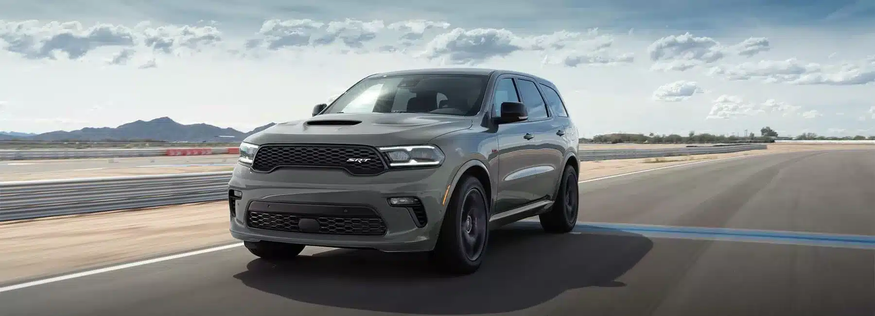 Gray SUV driving on a racetrack under a partly cloudy sky, showcasing its sporty design and performance.