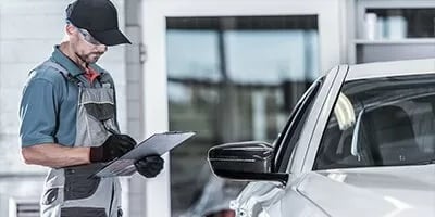 Mechanic in uniform inspects a car while writing on a clipboard in a garage setting.