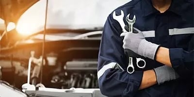 Mechanic in uniform holding wrenches near an open car hood, symbolizing auto repair services.